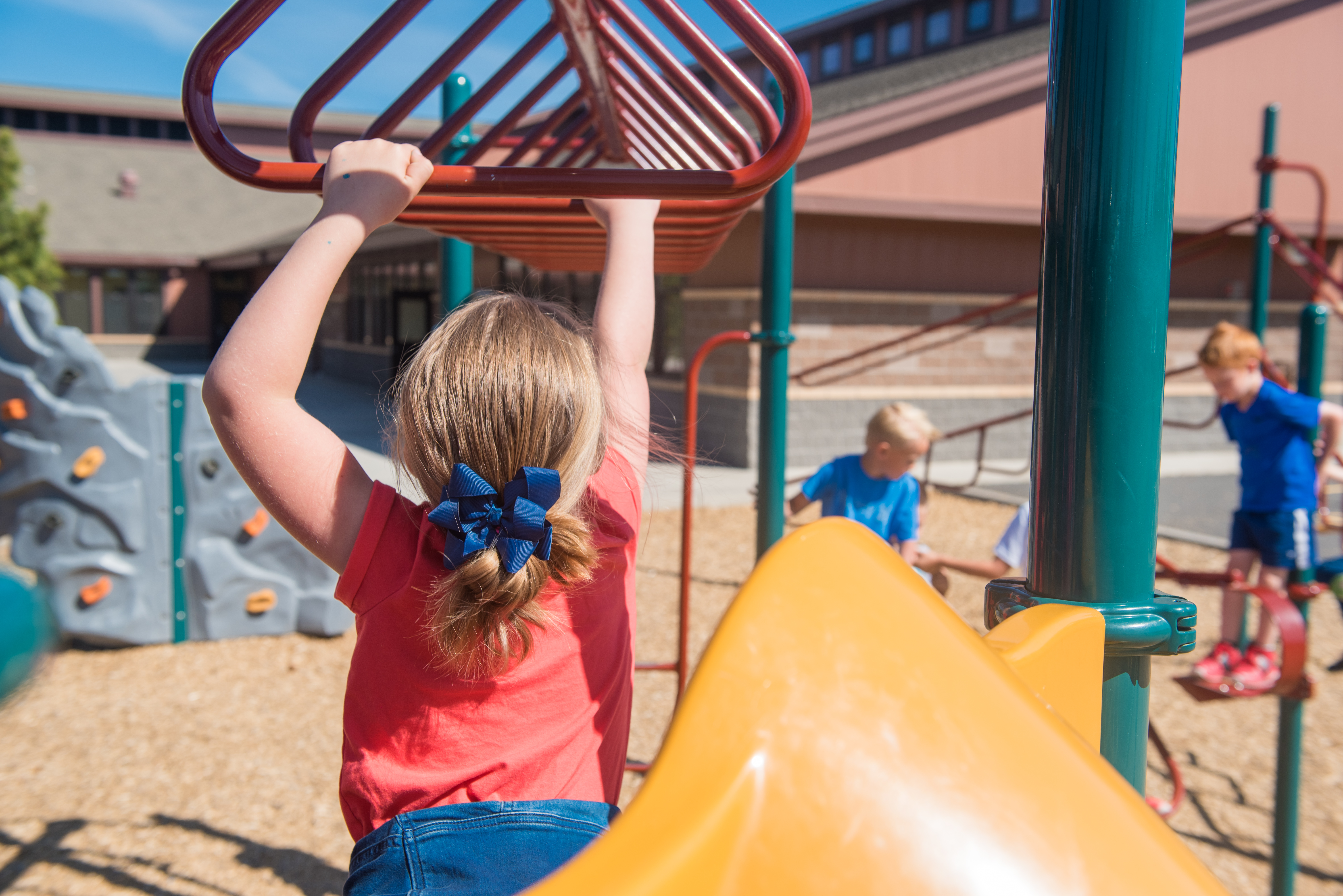 Elementary-age girl holding on to monkey bars in playground. Photo taken behind her. She's wearing a red shirt and has dark blond hair tied up with a dark blue bow.