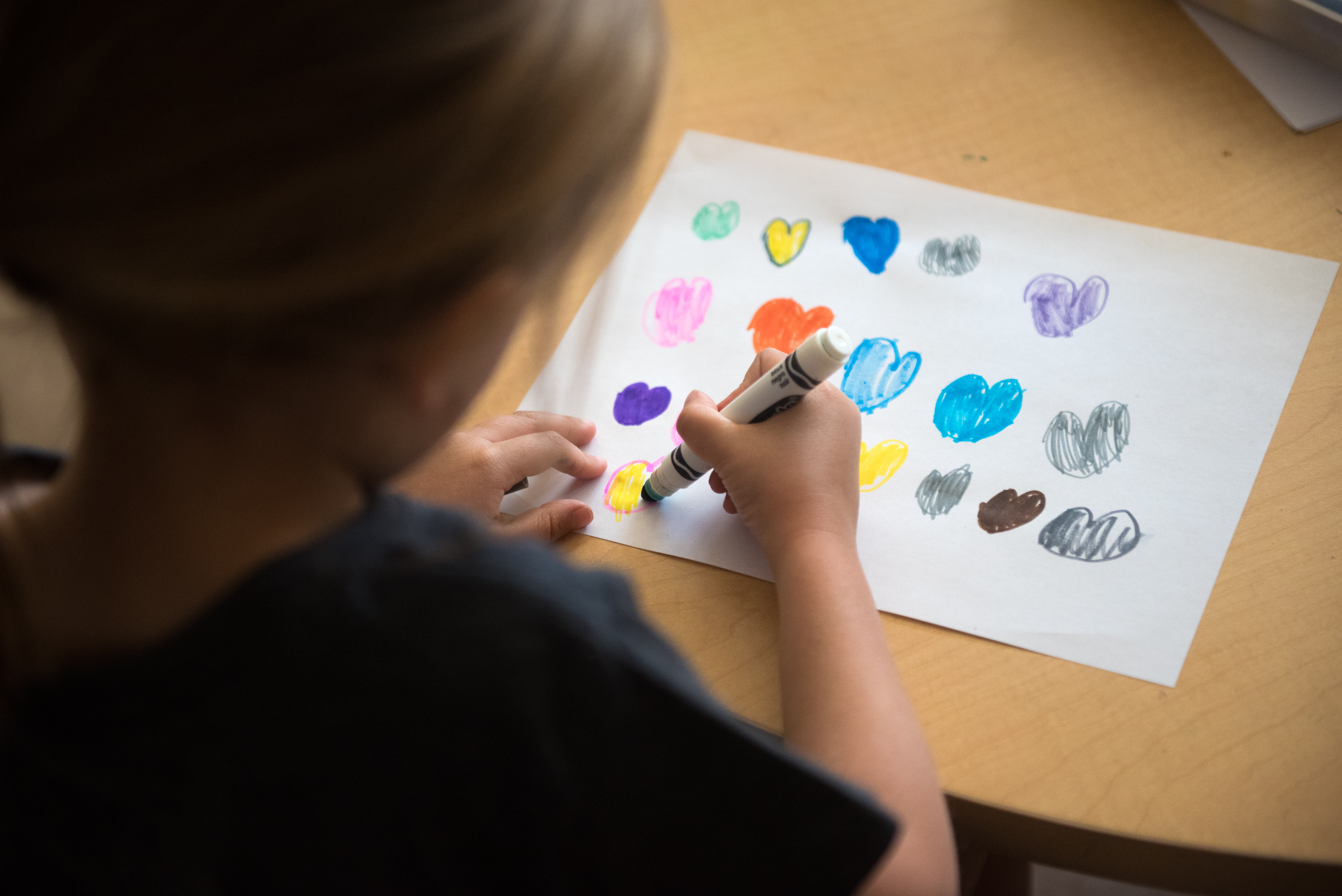 Looking over child's shoulder while she draws different colored hearts on a white sheet of paper.
