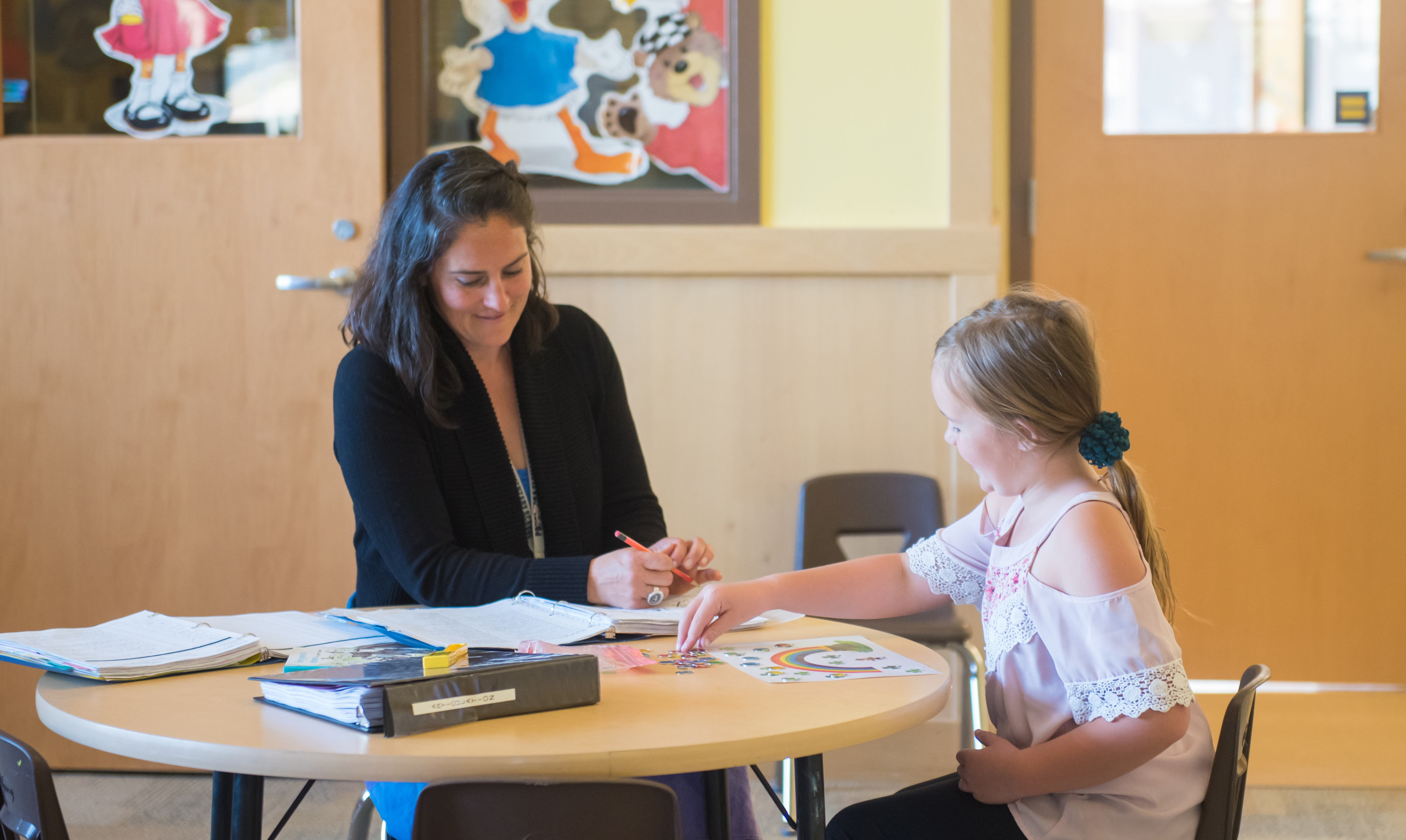 Adult woman with shoulder-length dark, straight hair sits at small round table with young girl and helps her with paperwork. Both are smiling and have arms out and leaning on work table. Woman is in dark sweater and girl is wearing white blouse.